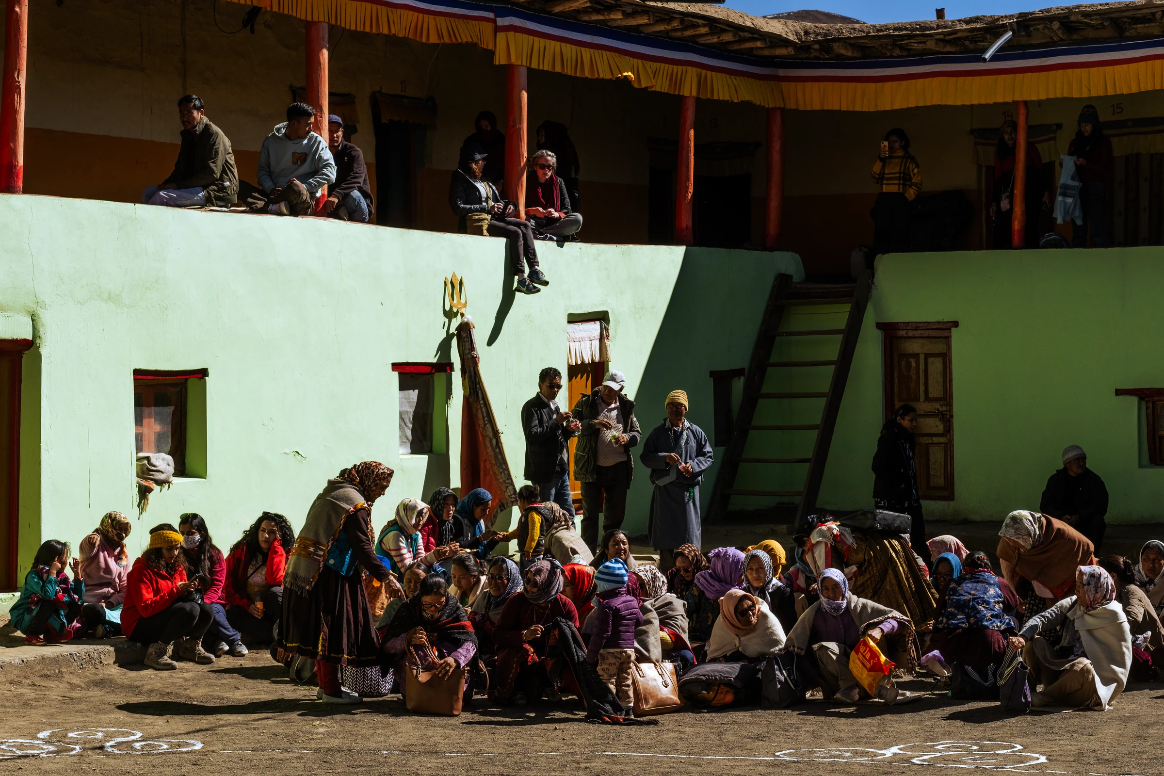 Spectators sit and stand on two levels around a pale green monastery courtyard, with sunlight and deep shadows crossing the walls.