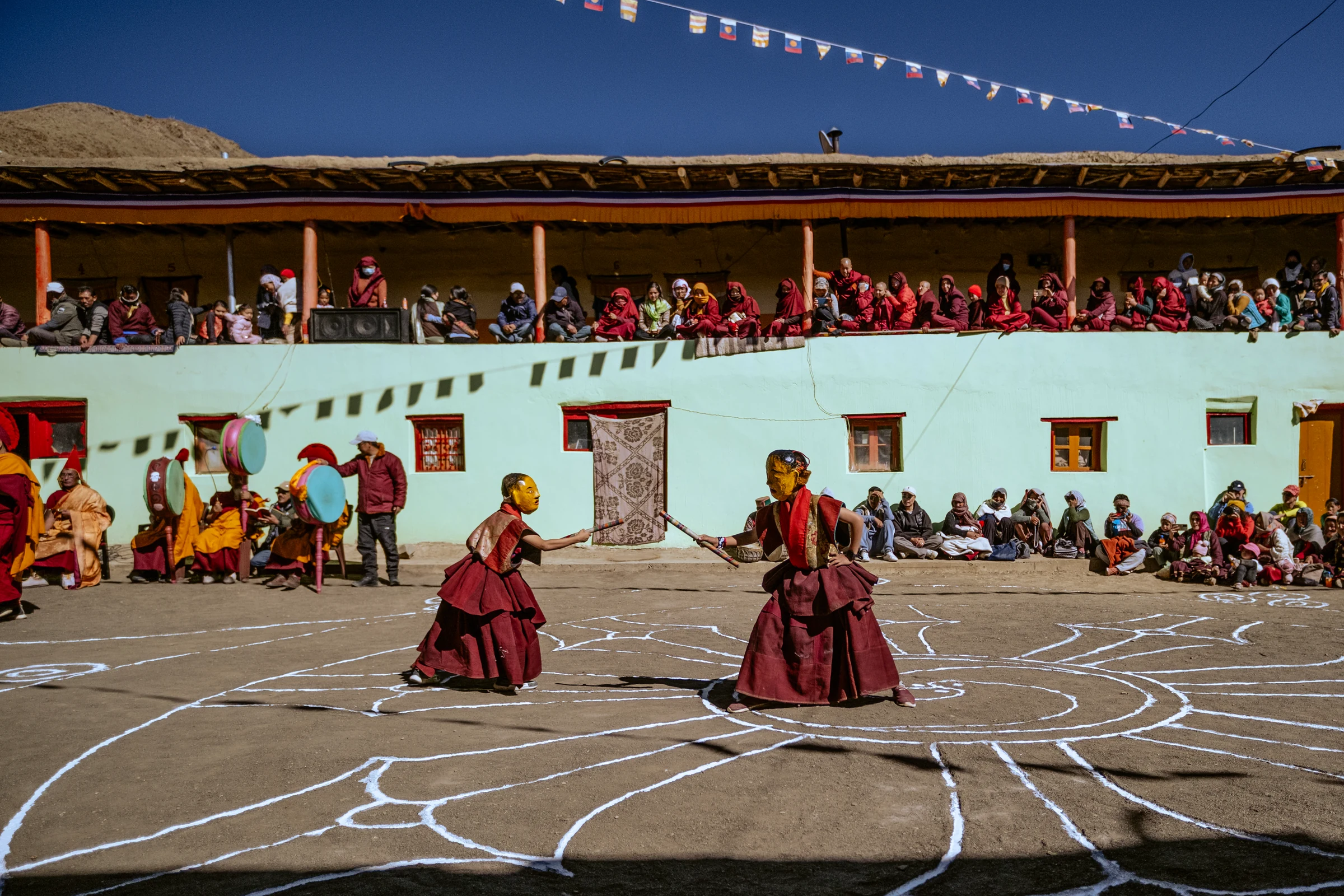 Two performers wearing gold-colored face masks and maroon robes face one another with short striped sticks, while musicians and spectators watch around the courtyard.