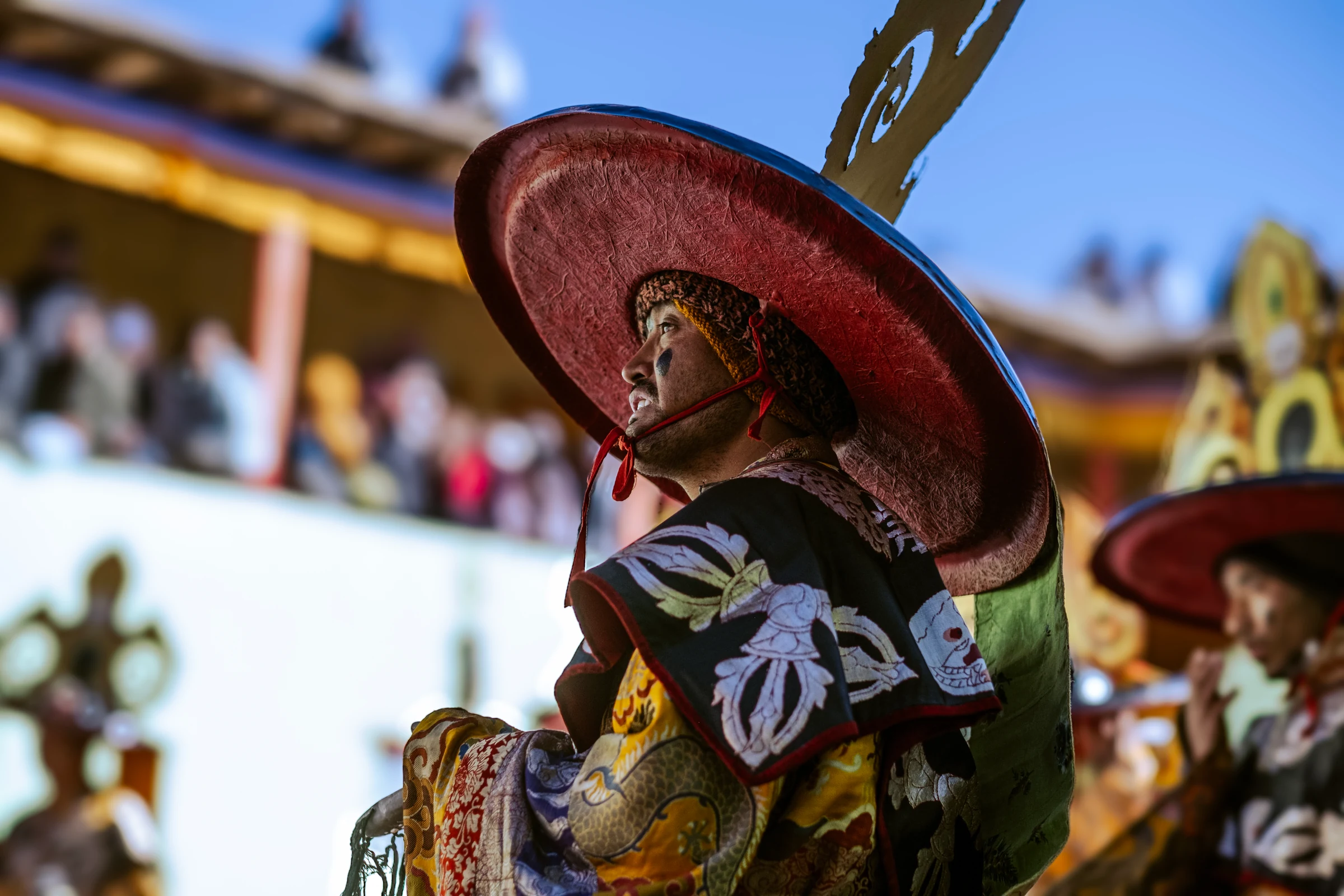 Close side view of a dancer beneath a broad red ceremonial hat, wearing embroidered black and gold robes, with the monastery softly blurred behind.