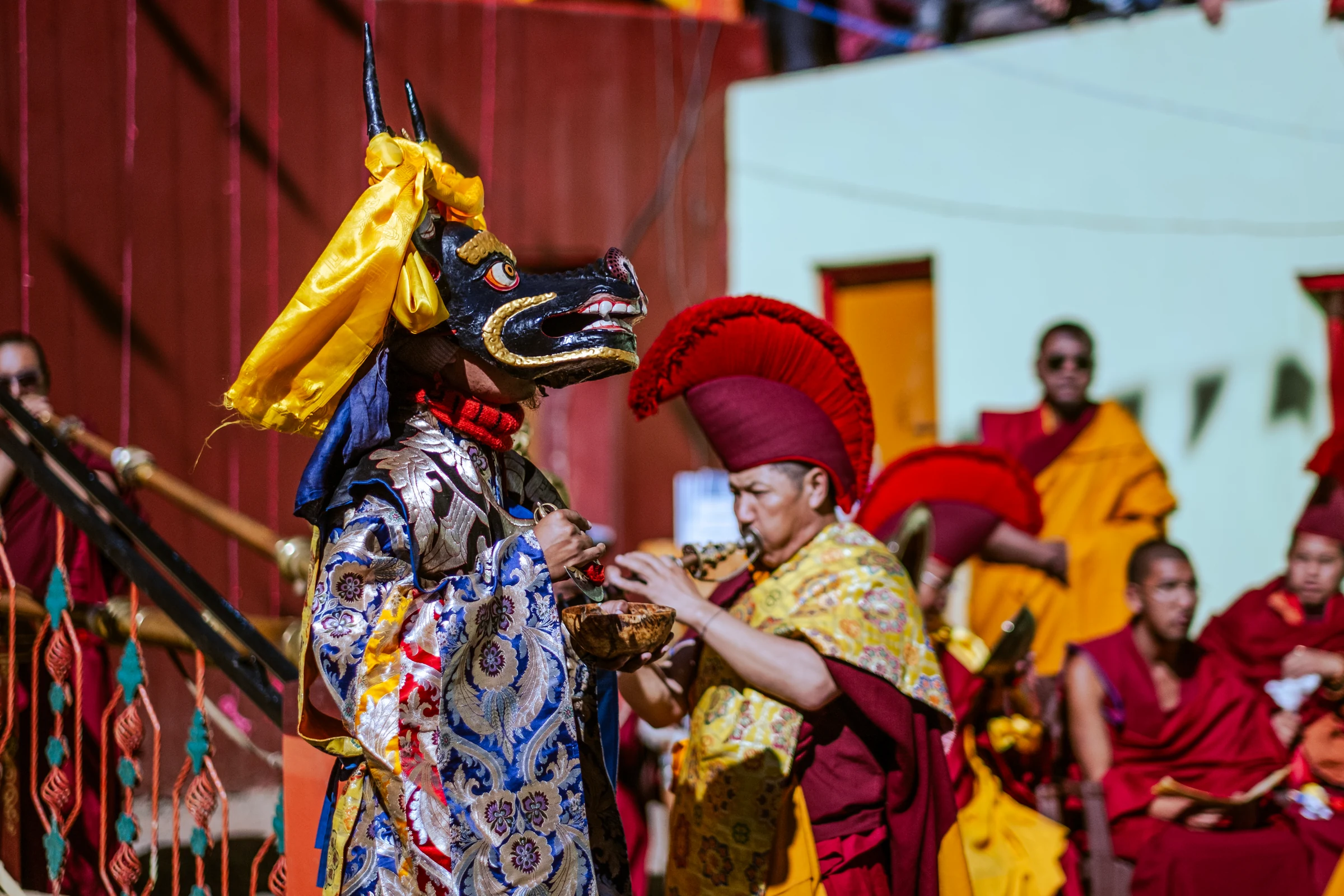A horned masked performer holds a bowl near a monk playing a wind instrument, with other monks in red and yellow robes seated behind.