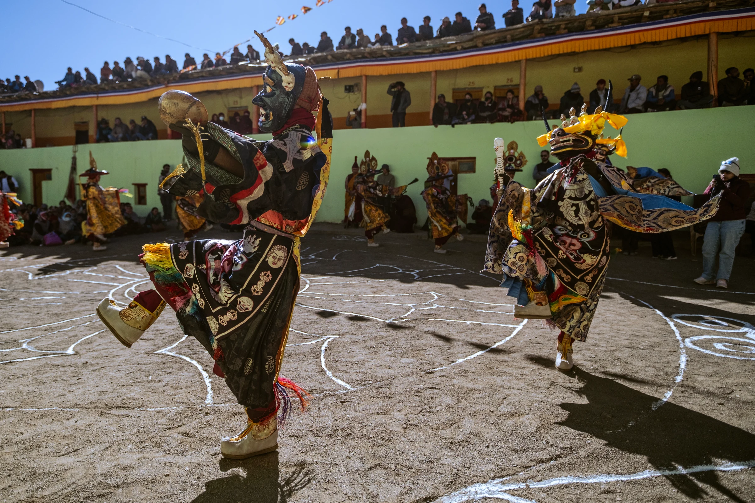 Two masked dancers lift their legs during a courtyard dance. Their patterned robes and ribbons move in sunlight, with spectators lining the balcony.