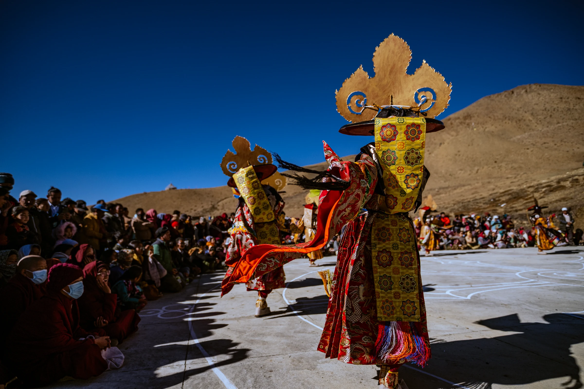 Two elaborately dressed dancers with broad ceremonial hats extend their red sleeves in an open courtyard bordered by spectators and a bare hillside.