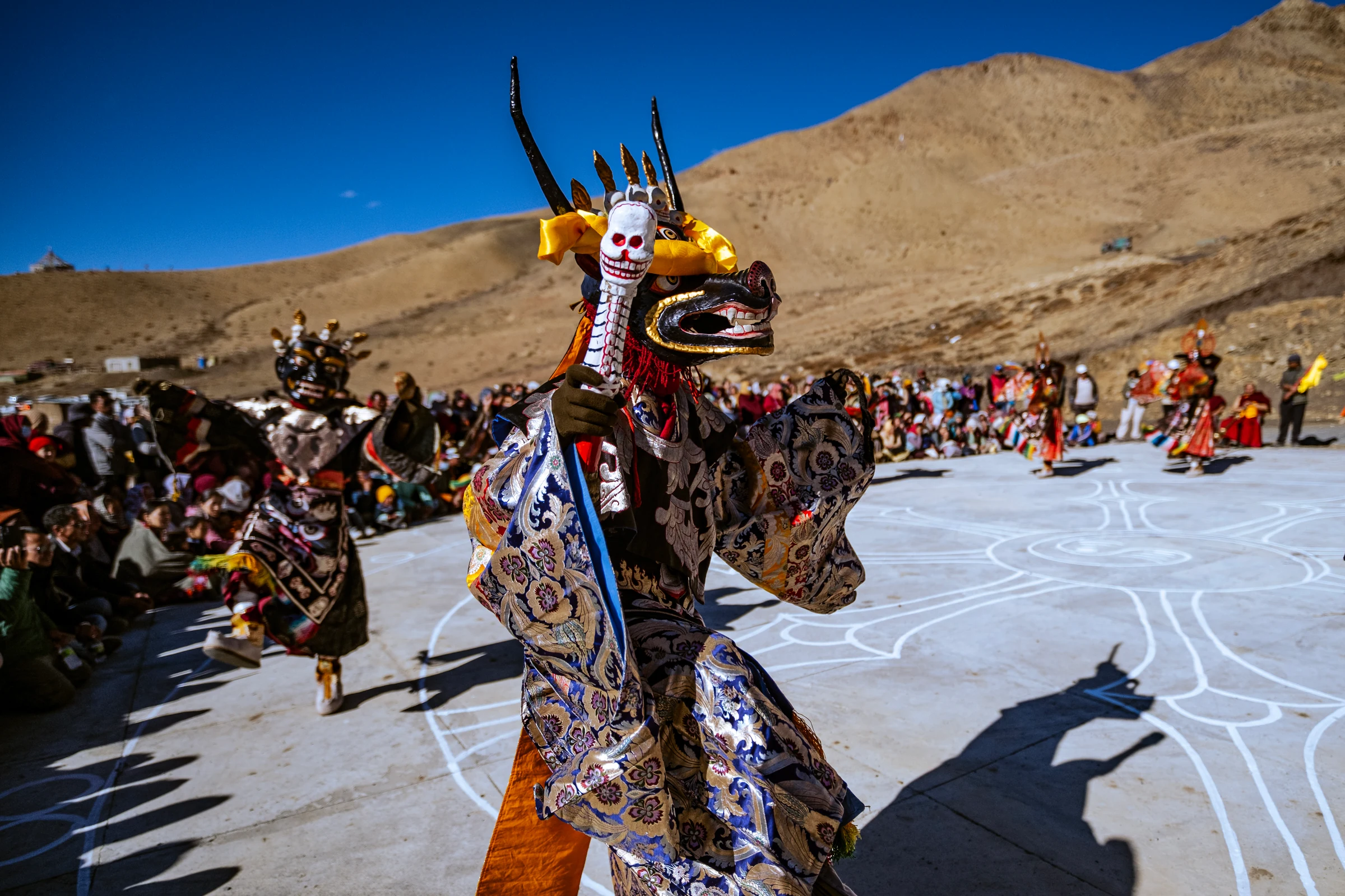 A dancer in a horned black mask, yellow ribbons and blue floral robes holds a white skull-topped staff in Komic’s courtyard, with spectators and dry hills behind.