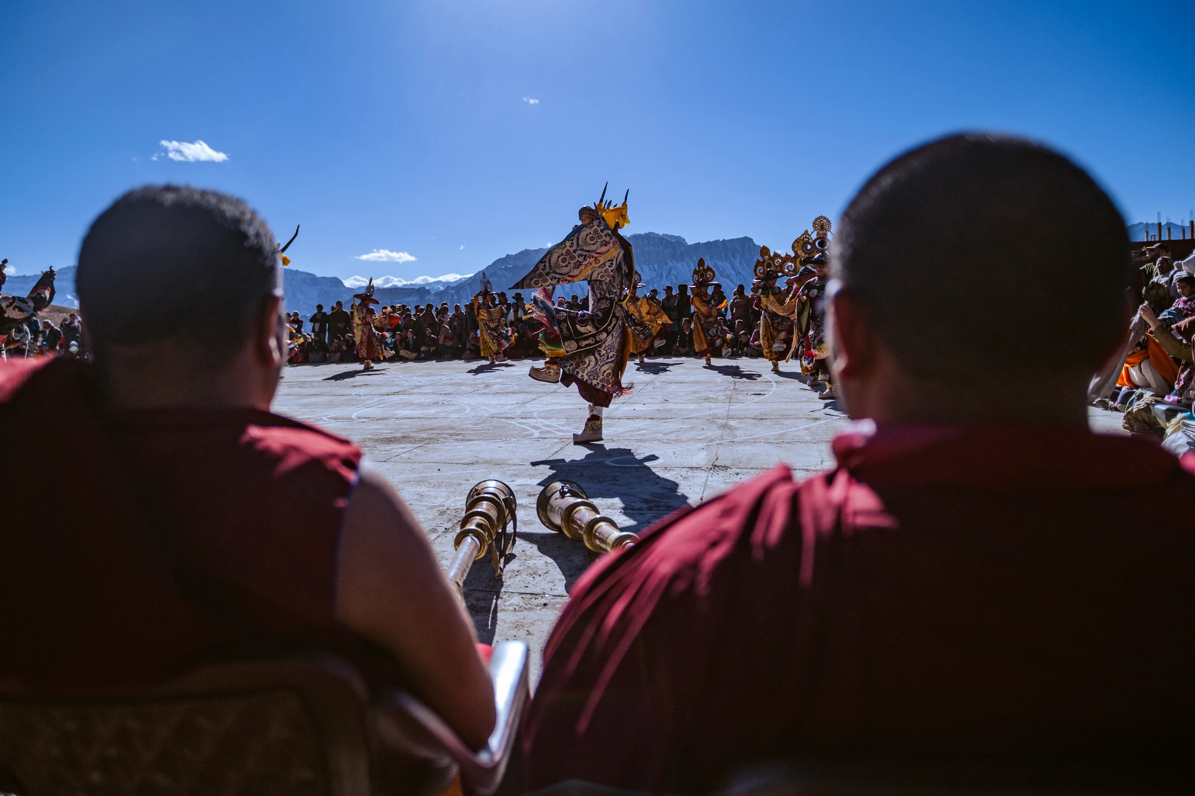 Seen between two seated monks in the foreground, a masked dancer lifts one leg beyond two long metal horns, with mountains and a crowd in the distance.