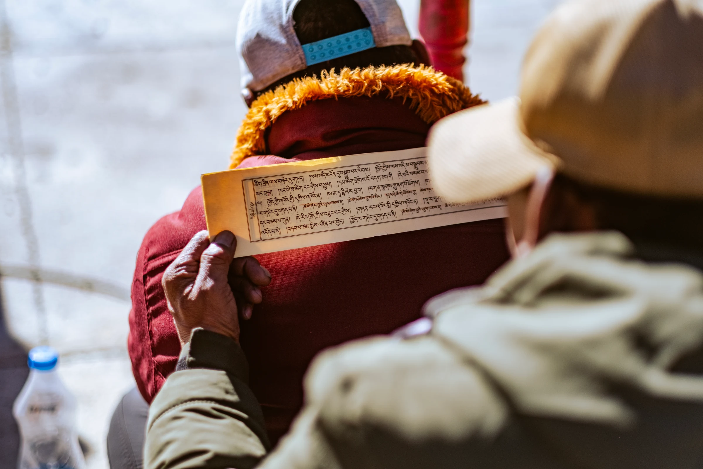 A spectator seen from behind holds a narrow page of Tibetan script beside another person wrapped in maroon fabric.