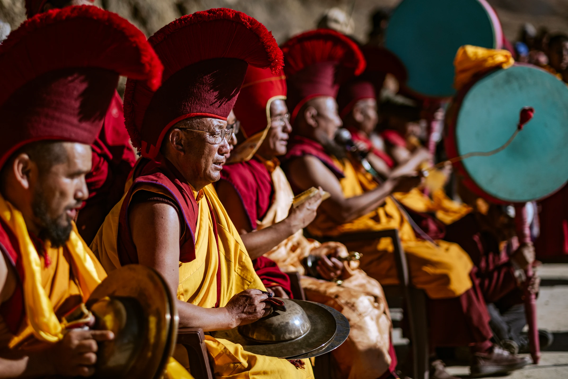 A row of monks in red ceremonial hats and yellow robes plays cymbals and drums. One monk’s face is in focus, with turquoise drums to the right.