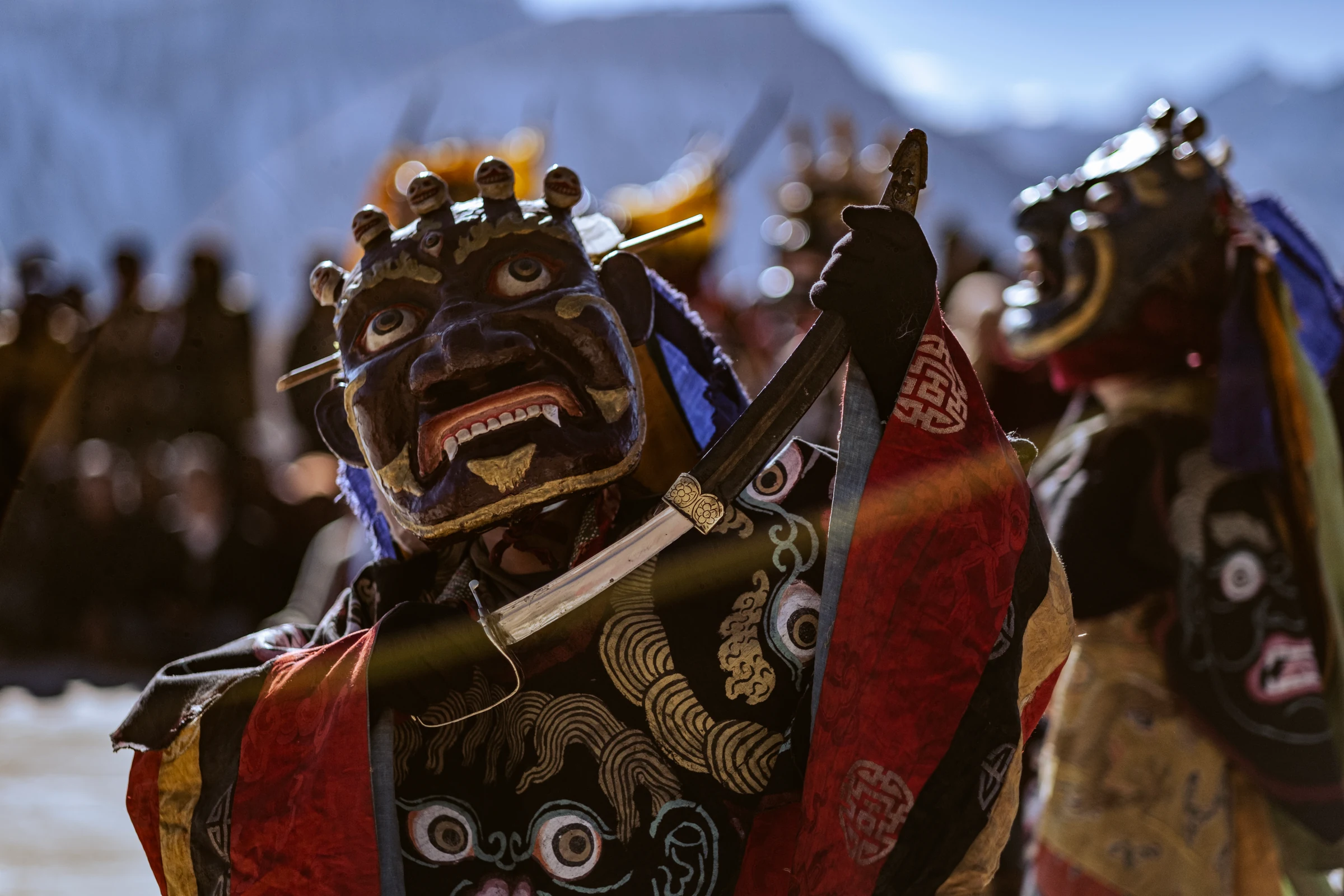 Close-up of a dark blue ceremonial mask with wide eyes, visible teeth and small skull shapes above it. An embroidered robe and a diagonal ceremonial prop fill the foreground.