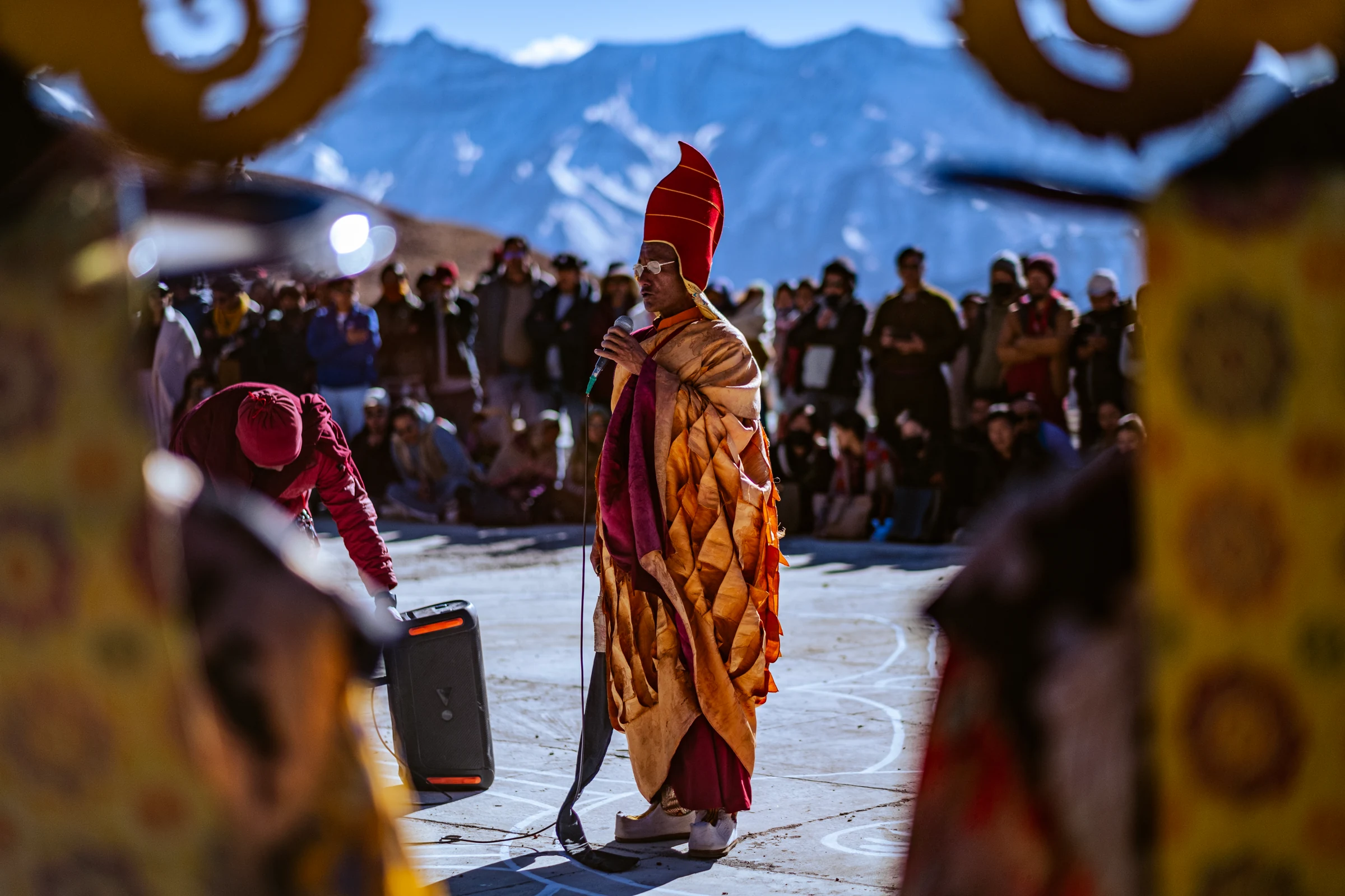 A monk in gold robes and a tall red hat speaks into a microphone in the courtyard, framed by blurred dancers’ costumes and mountains behind.
