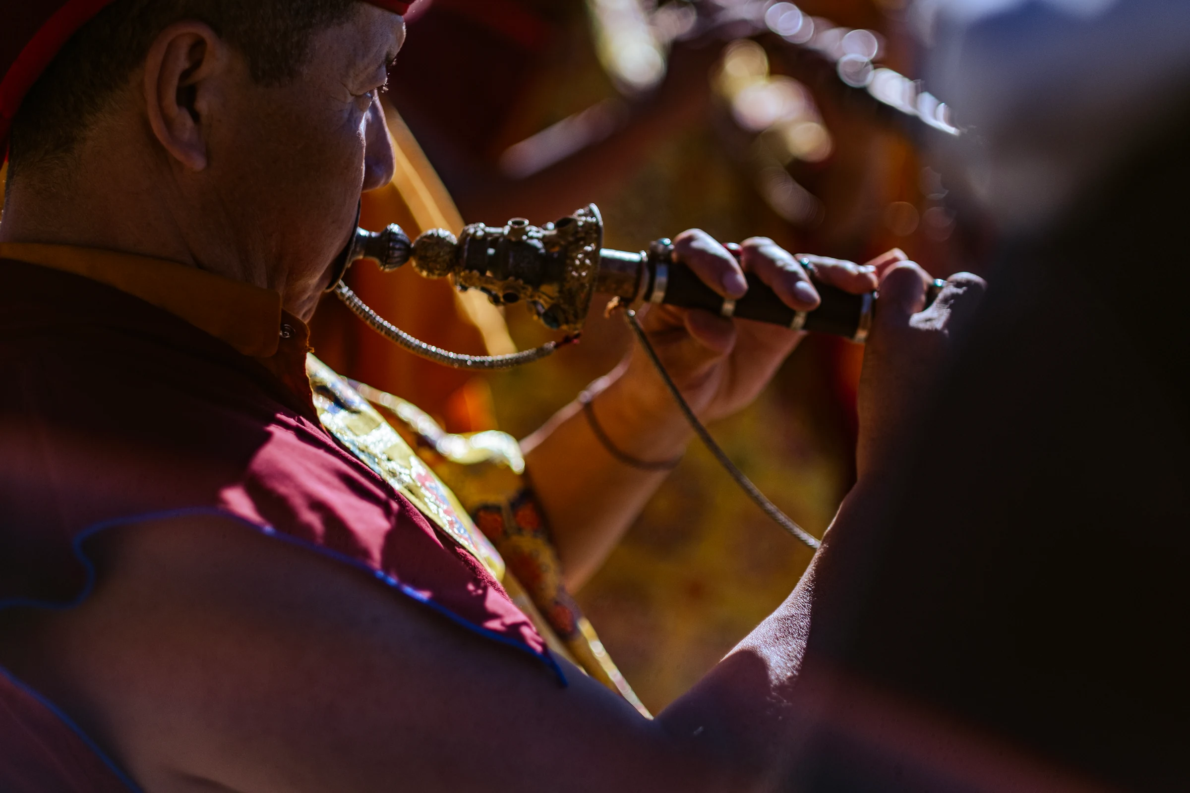 Close view of a monk’s face and hands as he plays an ornate wind instrument, with maroon robes and gold fabric in warm light.