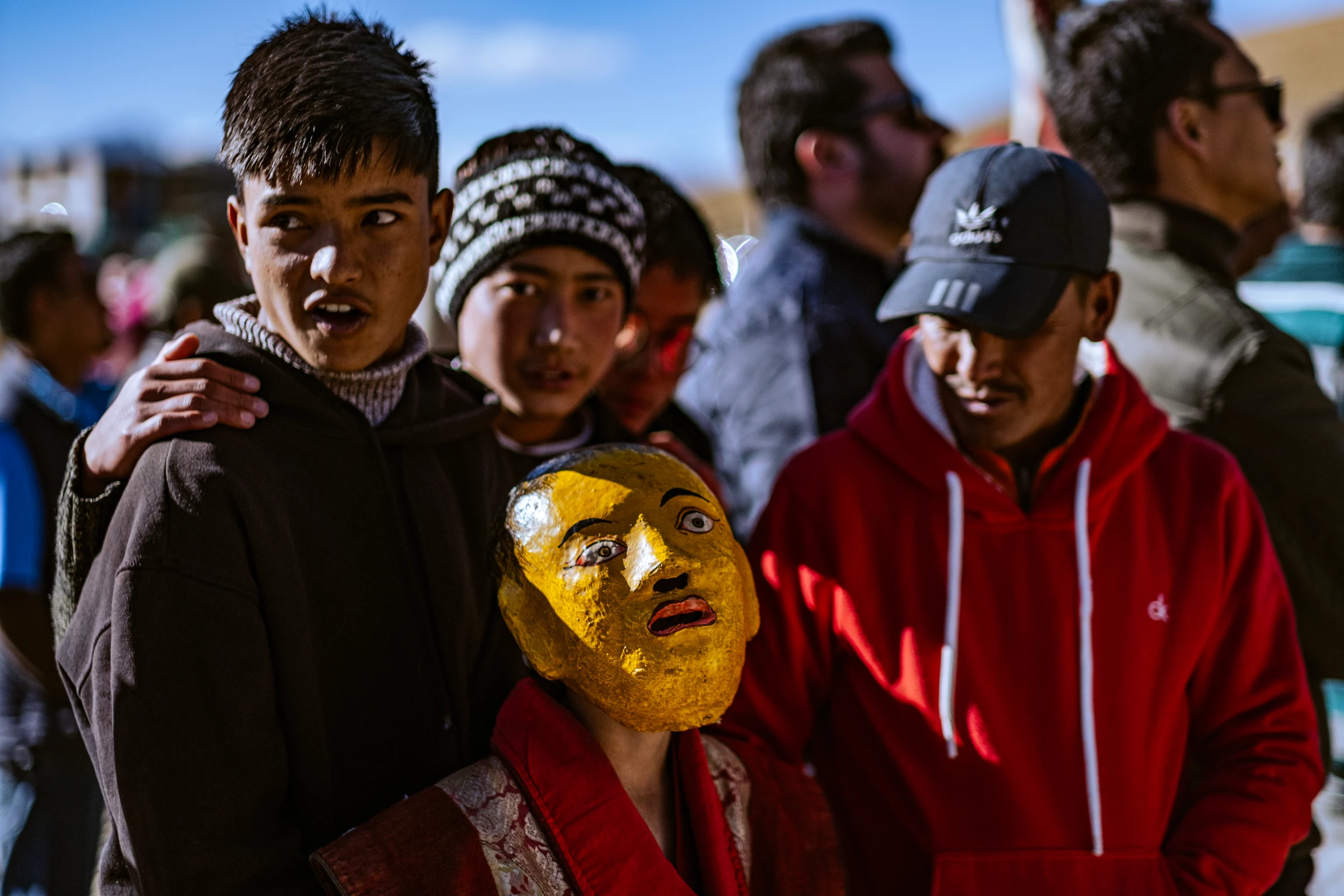 Young spectators stand close together beside a gold-colored face mask in the foreground, with a person in a red hoodie to the right.