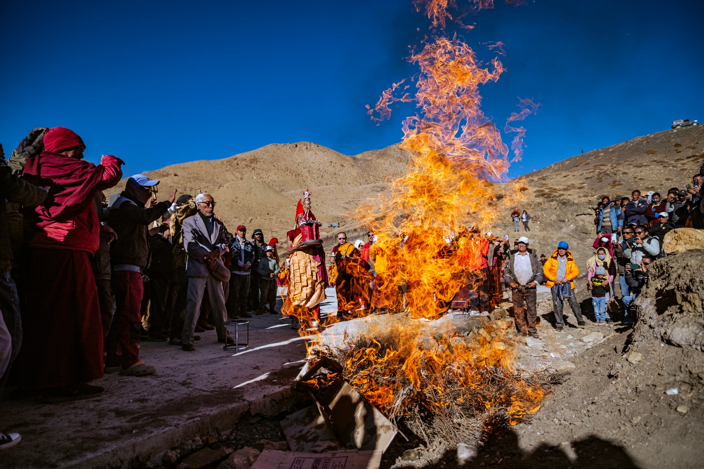 A tall orange flame rises from brushwood, surrounded by monks and spectators outdoors beneath bare hills and a deep blue sky.