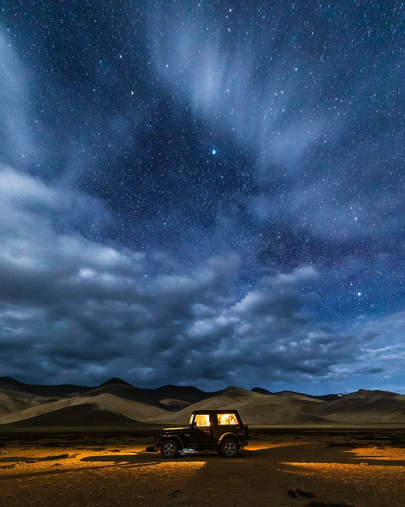 A vehicle lit from within on an open mountain plain under stars and streaks of cloud.