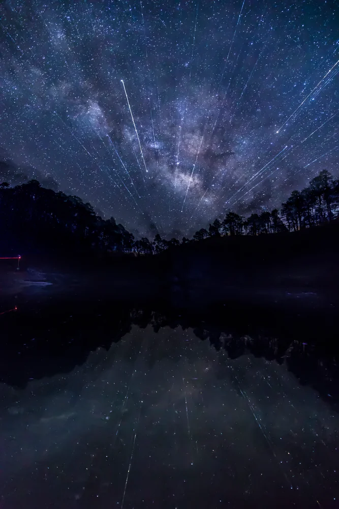 Bright streaks among stars above a tree-lined ridge, reflected in still water.