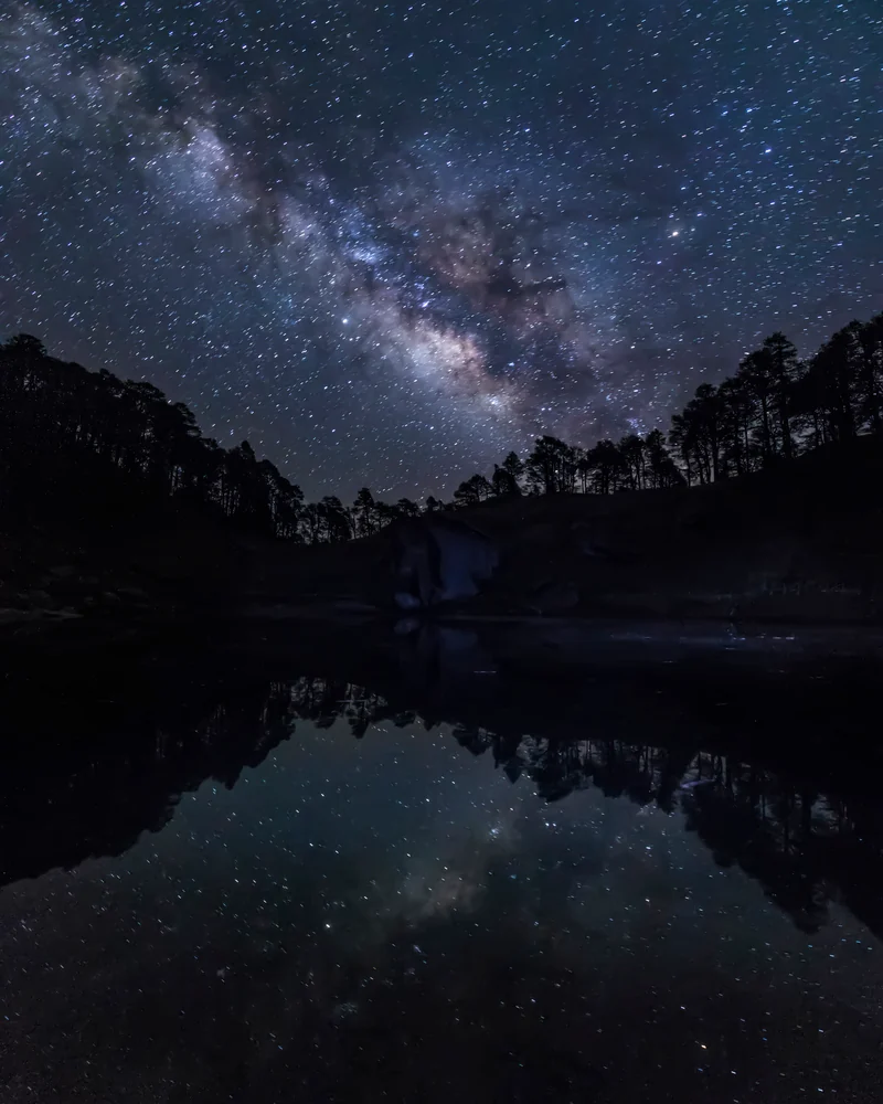 The Milky Way reflected in still water below a dark ridge lined with trees.