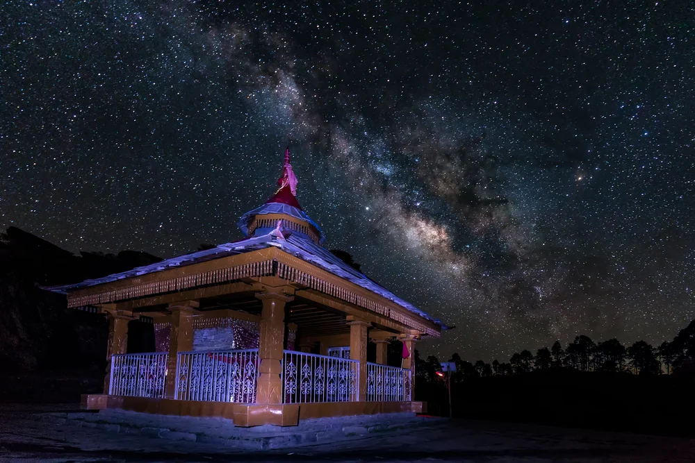 A small illuminated pavilion beneath the Milky Way and a sky filled with stars.