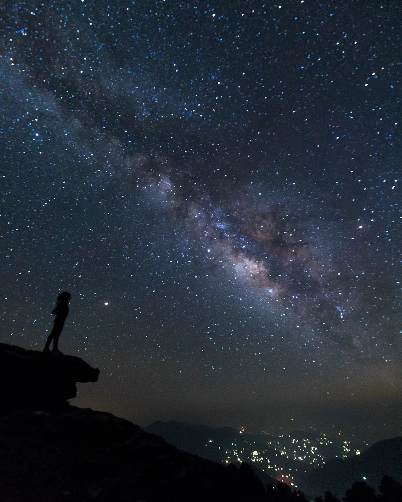 A person standing in silhouette on a rocky outcrop beneath the Milky Way, with distant lights below.