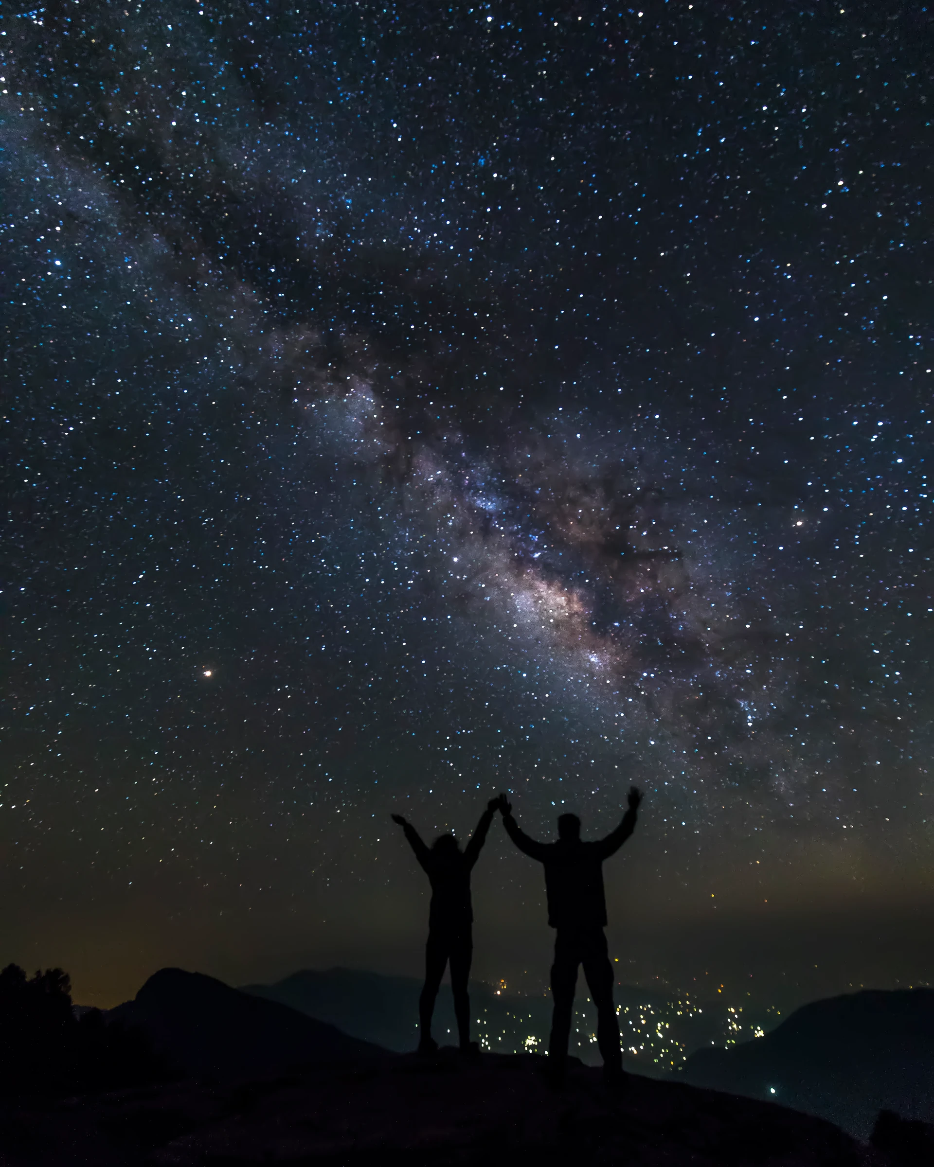 Two people standing with raised arms in silhouette beneath the Milky Way above distant lights.