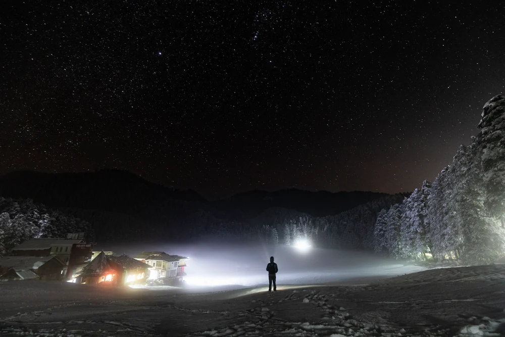 A person standing beneath a star-filled sky beside bright beams of light and snow-covered trees.