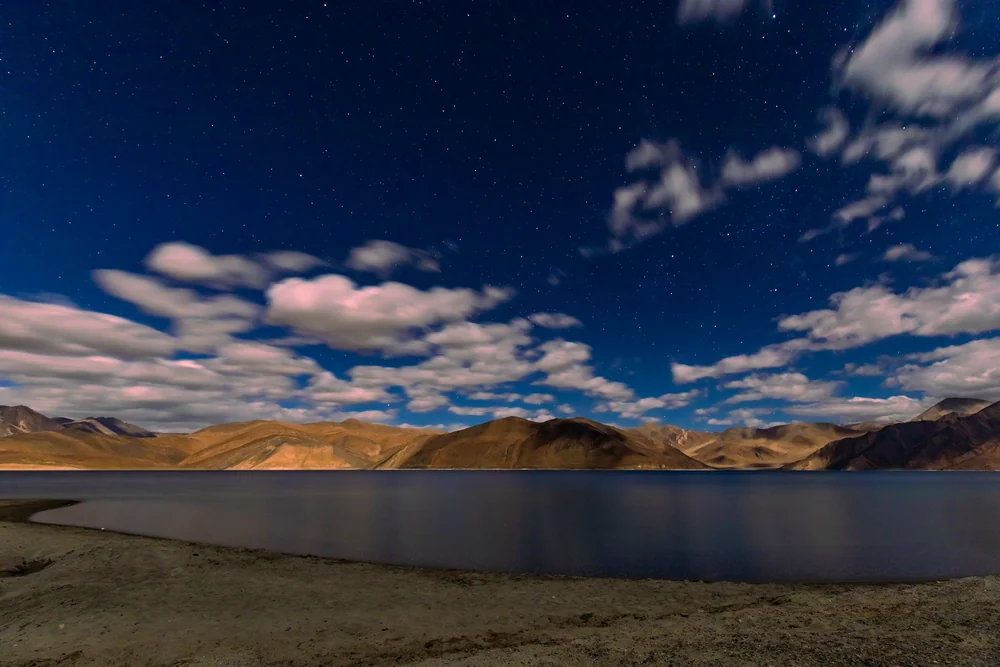 A mountain lake beneath a deep blue star-filled sky and pale clouds, with the shoreline softly illuminated.
