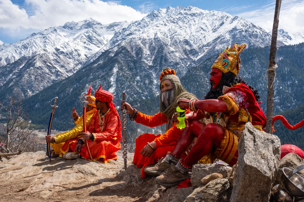 Costumed festival participants seated on rocks in front of snow-covered mountains.