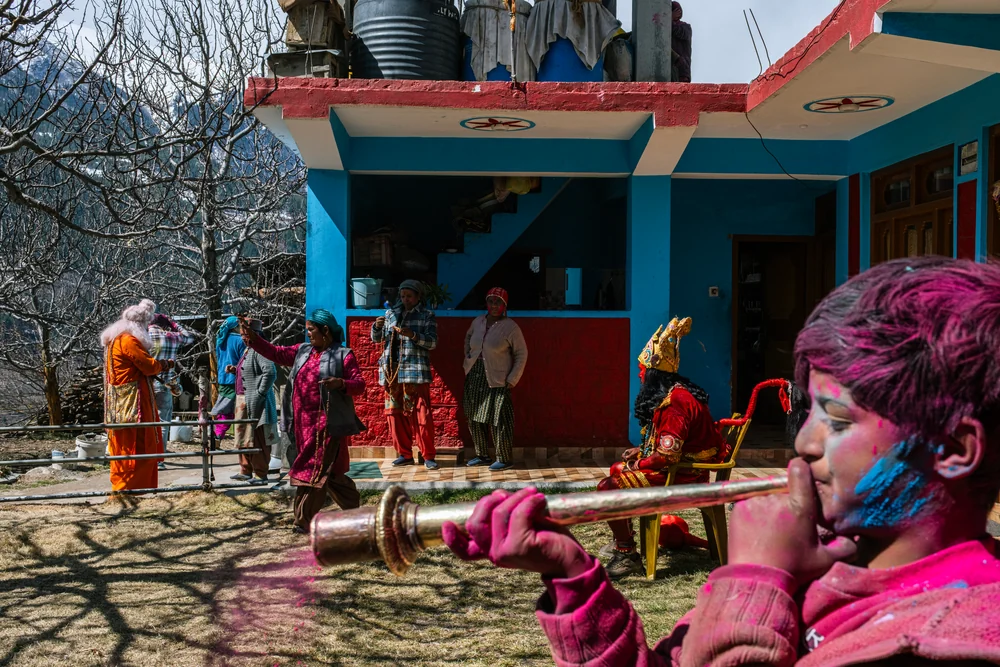 A musician with coloured powder on their face playing a brass wind instrument outside a brightly painted house.