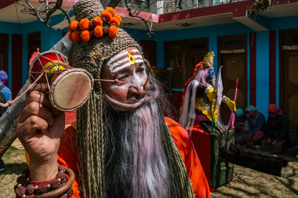 A festival participant in orange robes, a long grey beard and face paint holding a small hand drum.