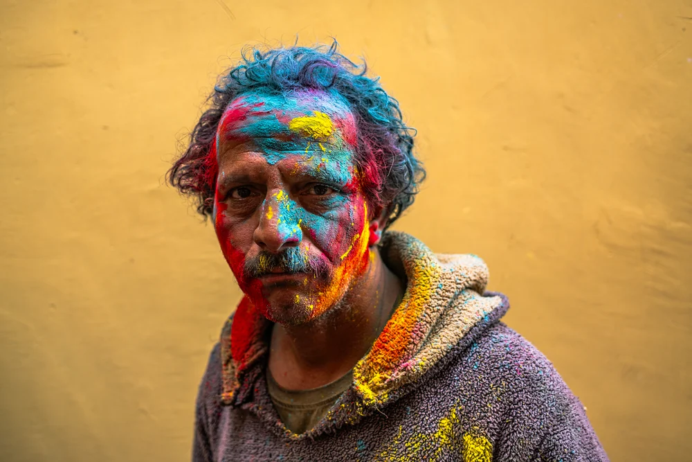 A man with blue, red and yellow powder on his face standing against a warm yellow wall.