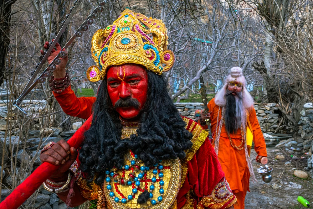 A participant wearing a red mask and an ornate gold crown walking beneath bare branches, with another costumed figure behind.
