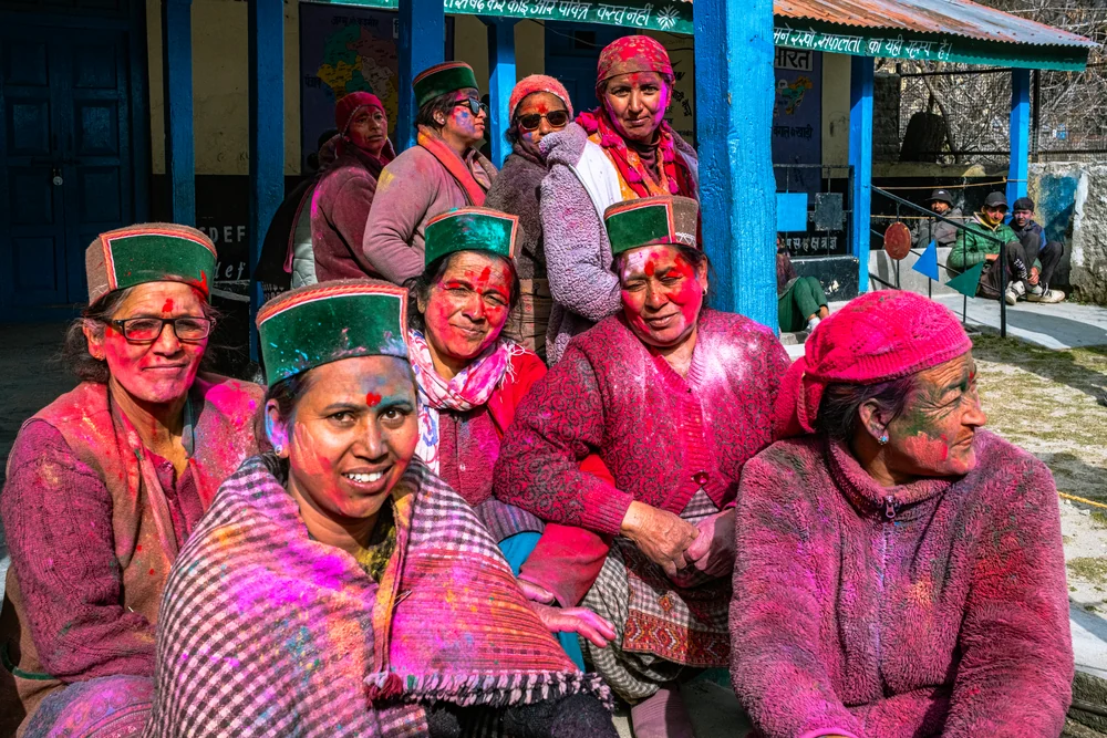 A group of smiling women with coloured powder on their faces sitting outside a blue-painted building.