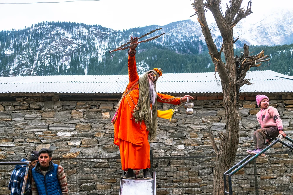 A festival participant in orange robes balancing above a stone wall with a raised staff and snowy hills behind.