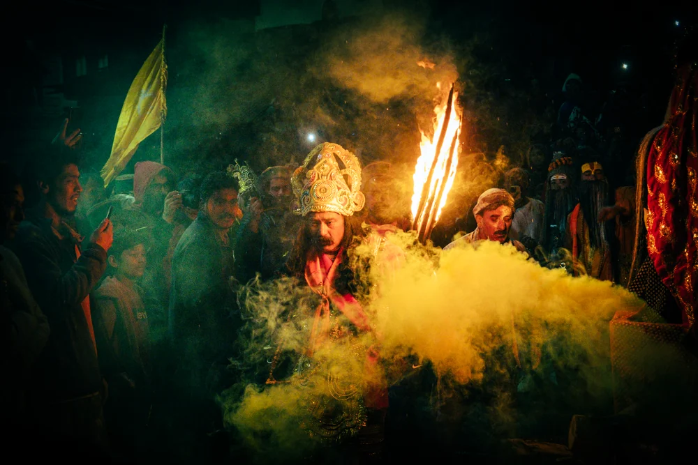 A crowned festival participant holding flaming torches amid a cloud of yellow powder and a nighttime crowd.
