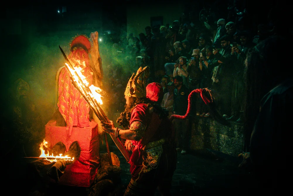 A costumed participant kneeling with flaming torches in front of another figure as a crowd watches at night.