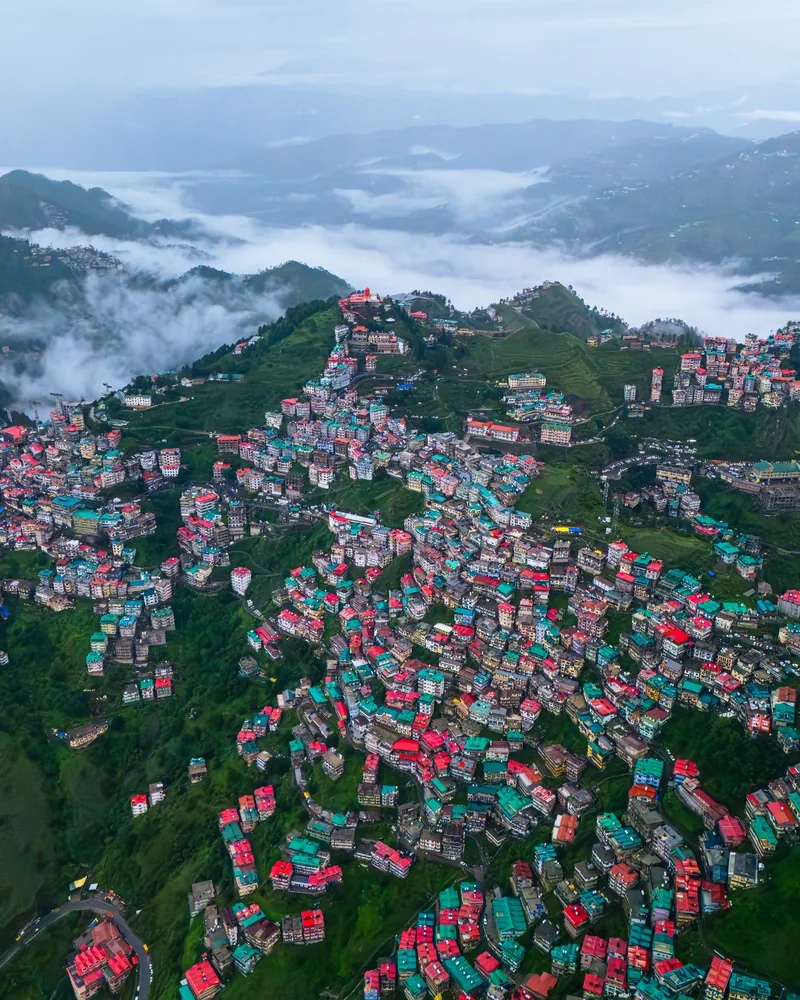 An aerial view of colourful buildings across green hills, with clouds filling the valley.