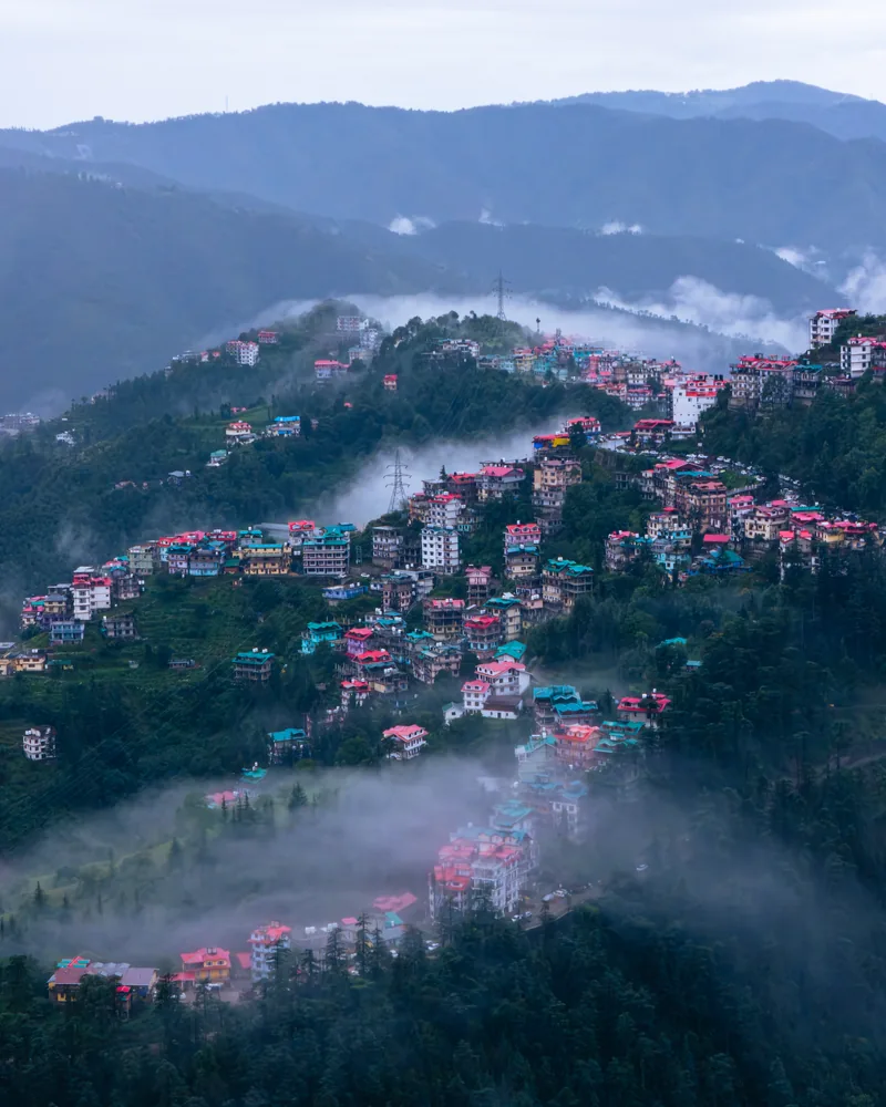 Rows of hillside houses framed by dark wooded slopes and drifting mist.