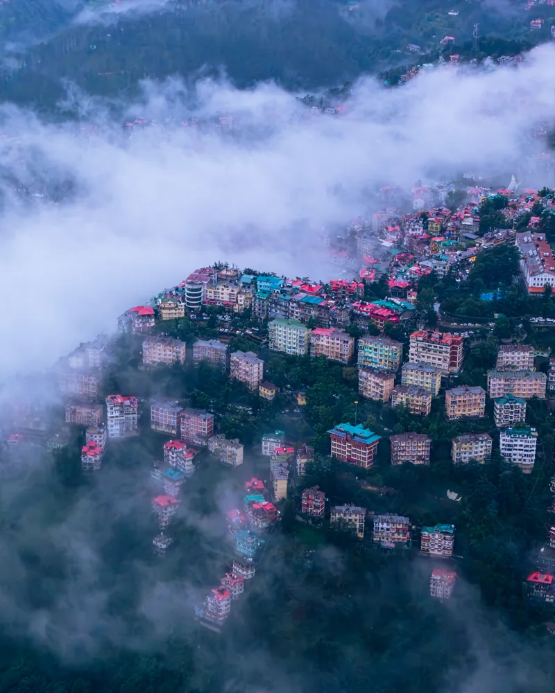 Buildings with red and green roofs appearing through clouds above a wooded hillside.