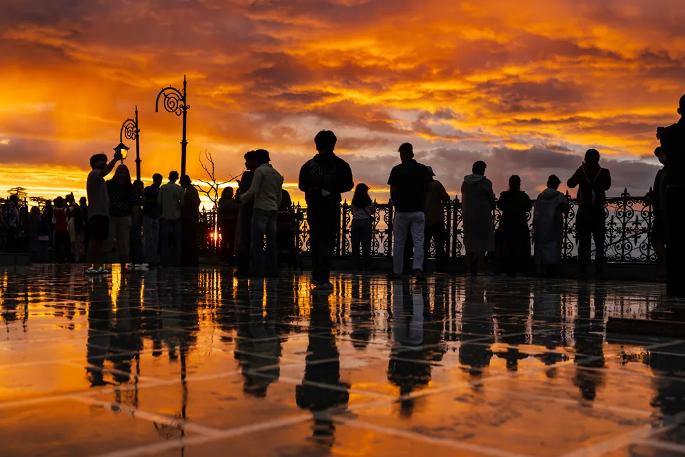 People silhouetted against an orange sky, reflected in rainwater on a promenade.