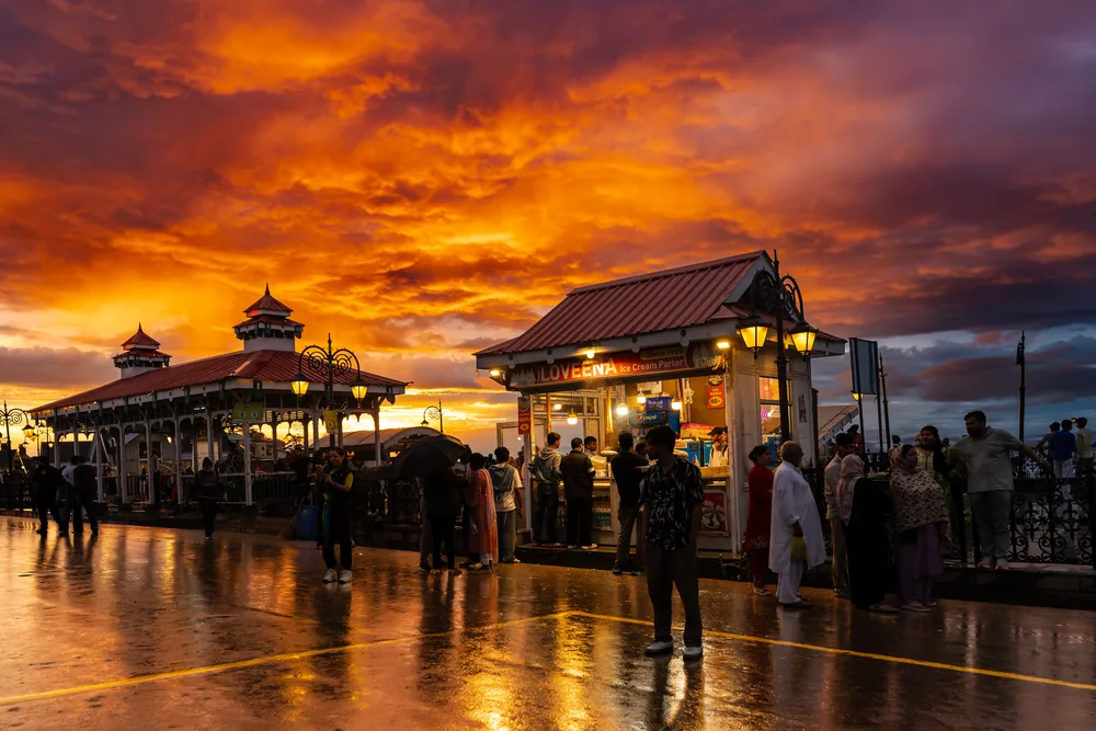 Illuminated pavilions and people on a wet promenade beneath a fiery orange sky.