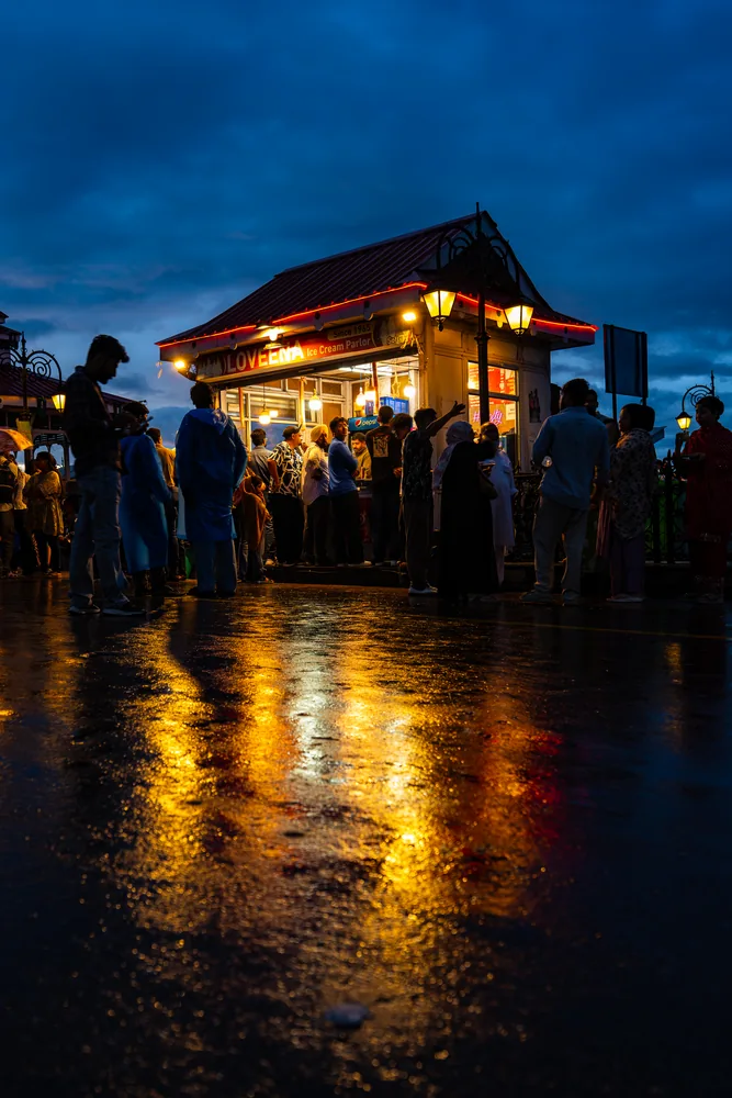 A lit pavilion and people reflected in wet paving under a deep blue evening sky.