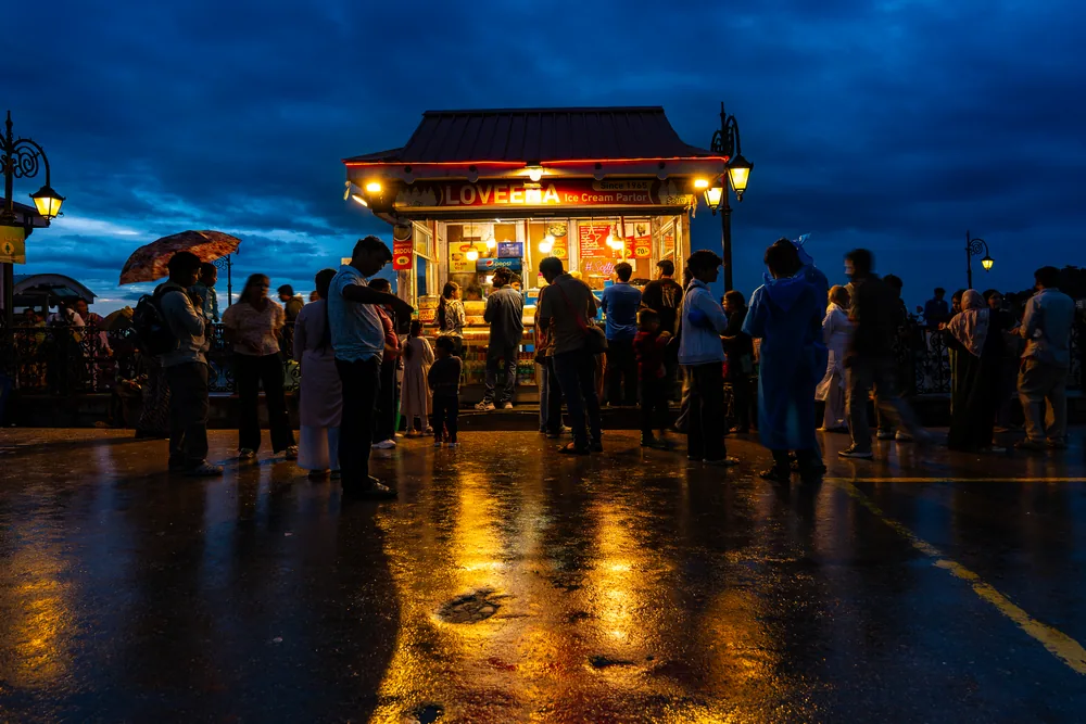 People with umbrellas outside an illuminated pavilion, reflected in wet paving at dusk.