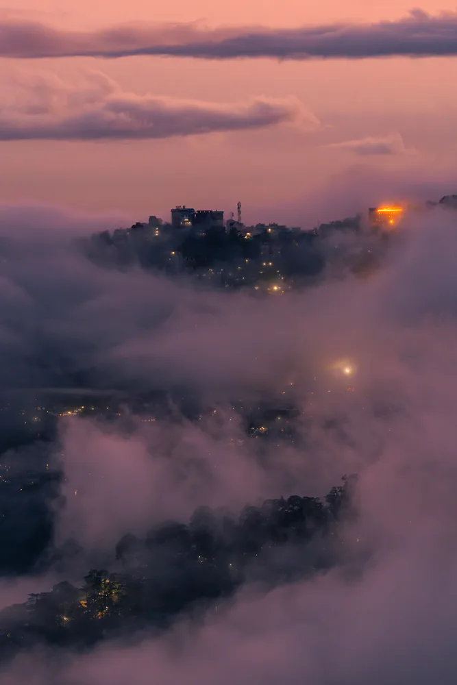 Clouds rolling around a wooded hilltop with a glowing light beneath a pink evening sky.